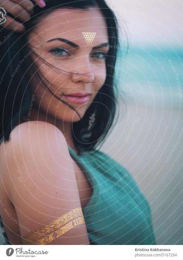 Outdoor fashion portrait of beautiful brunette lady at beach with flash tattoos on hands, back and forehead. Gypsy boho style. Woman in long green dress looking at camera.