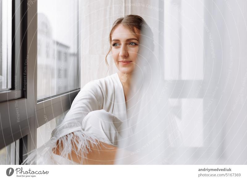 Bride s morning. Bride drinking champagne in the peignoir. young woman is sitting on a large window in a hotel room in bathroom. Beautiful girl in white wedding robe. wedding day
