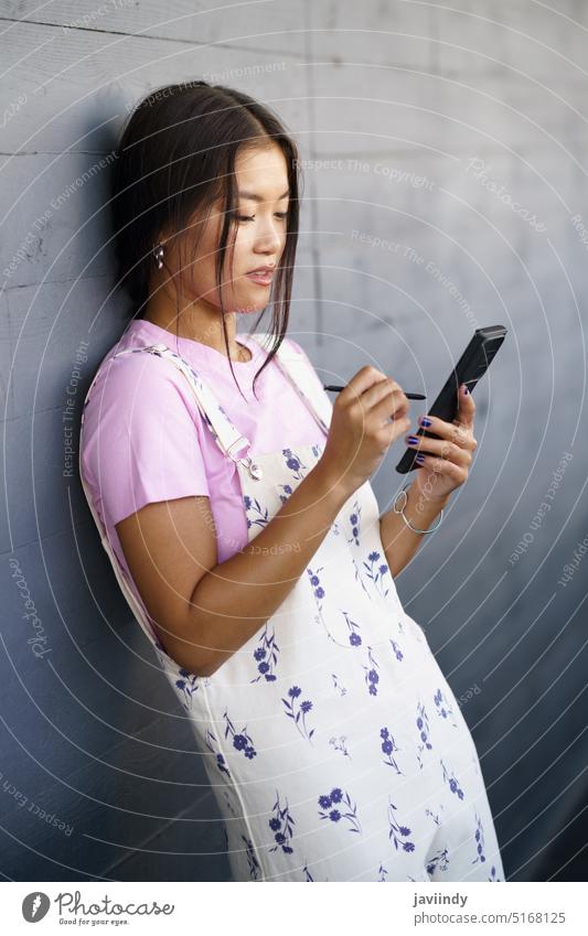 Asian female leaning on wall and using cellphone woman smartphone stylus style news street social media young ethnic asian online gadget device browsing mobile