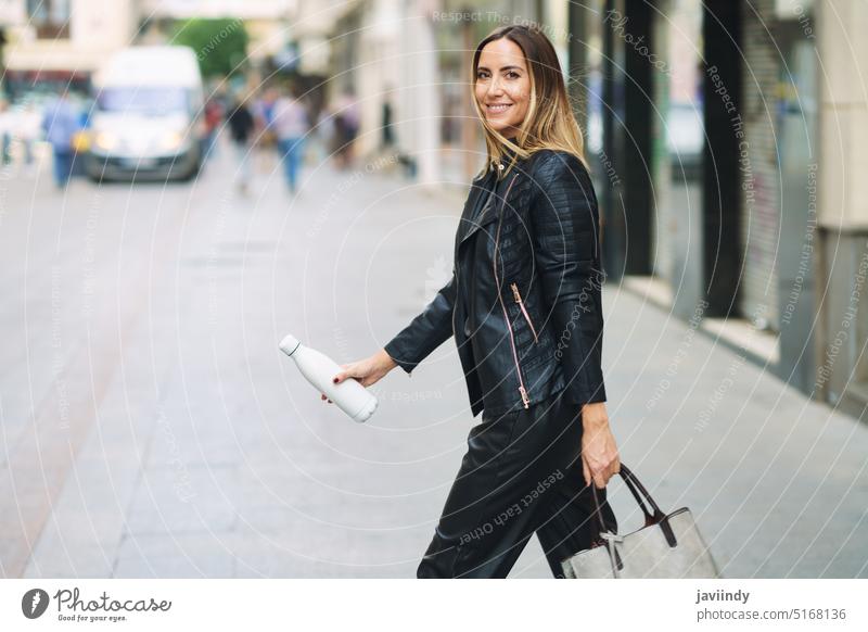 Smart casual lady with bag and bottle walking on street woman commute style urban town confident trendy contemporary modern zero waste leather jacket smile