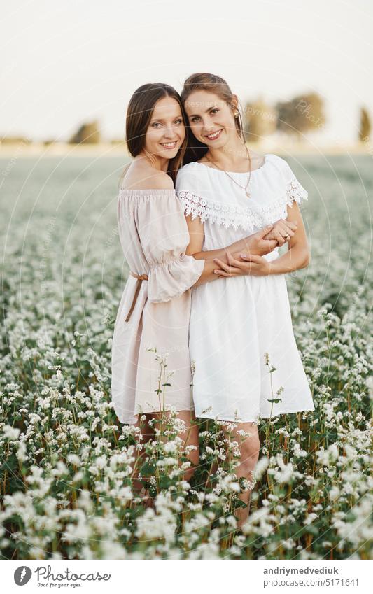 Portrait of beautiful young two women sisters in white dresses with long hair in a field with white wildflowers in summer. Rustic style, village. Tenderness, hugs, support.