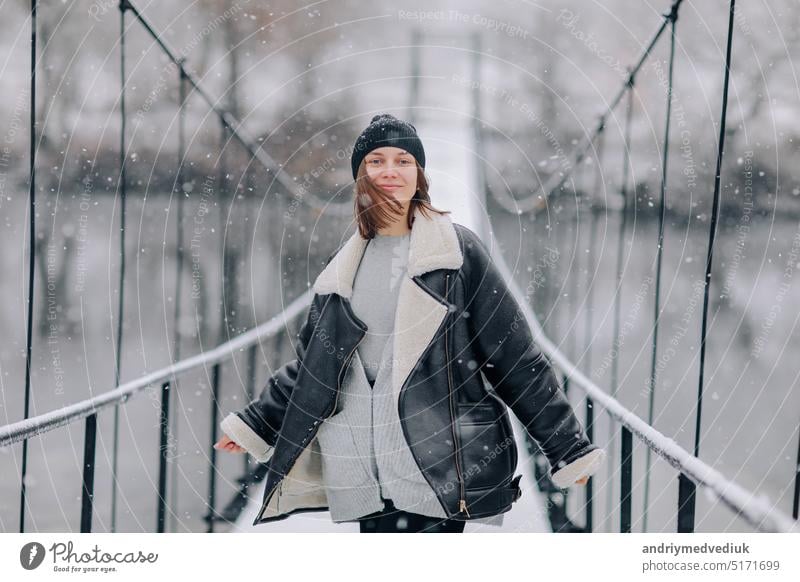 A woman walks over the river on a suspension bridge in winter day. Young girl in warm clothes stands on a wooden footbridge in cold snowy day nature young