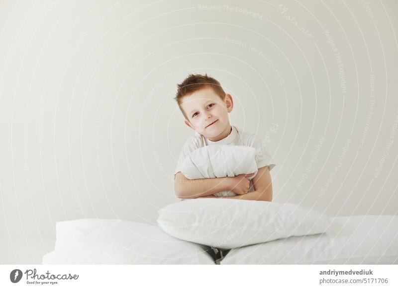 Portrait of casually dressed little boy is hugging white pillow, feeling tired after long full day, with cheerful smile, having rest sleep bed hand child person