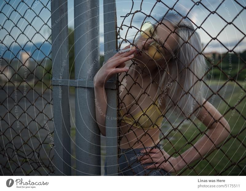 A gorgeous girl is wearing a yellow sexy bralette and smoking a cigarette. This rusty fence can’t get in her way. Like anything else. A pretty woman, with some bad habits, and a fashionable beauty.