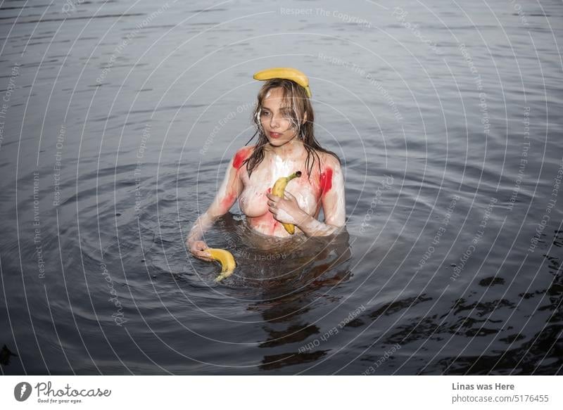 An image full of lust and sensual sexiness of a gorgeous female model. A pretty woman is taking a naked swim while being all coloured in body paint. An erotic picture with a wild and nude girl in a dark water. And bananas also.