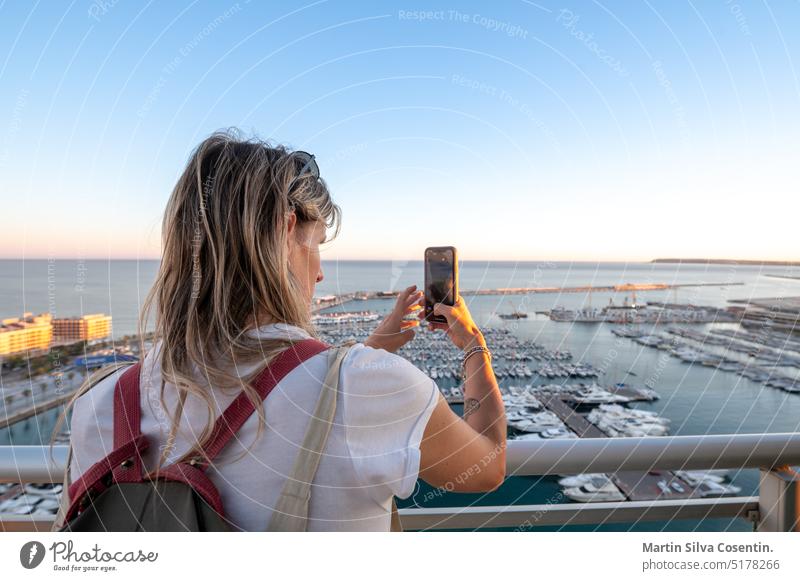 Woman takes a photo of the Port of Alicante from a hotel terrace in Spain adult alacant alicante architecture beach beautiful blue camera casual caucasian coast