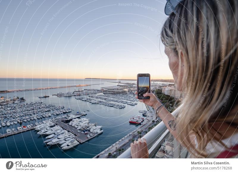 Woman takes a photo of the Port of Alicante from a hotel terrace in Spain. adult alacant alicante architecture beach beautiful blue camera casual caucasian
