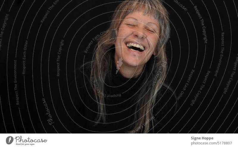 Woman with long gray hair and piercings wearing black sweater laughs heartily with closed eyes against black background Laughter grey Sweater Rolli Pircing Joy