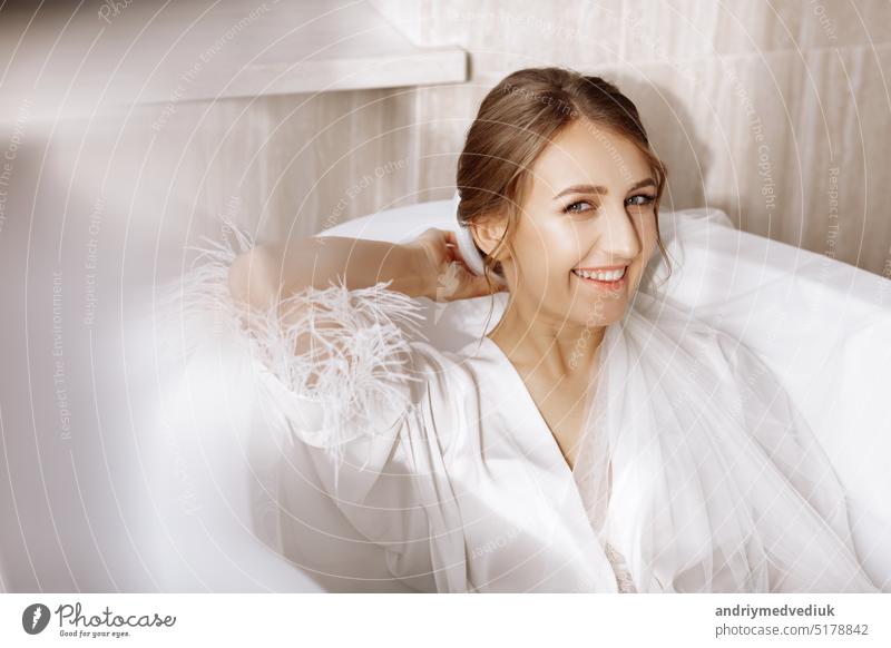 Morning of the bride. Happy beautiful young woman is wearing in a white long veil, robe and underwear sitting laughing and having fun in the bath on a white background, relax. wedding day