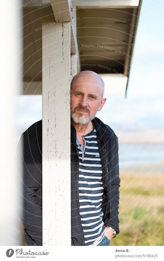 a man with a beard and a striped shirt leans casually behind a beam on the porch of a house by a body of water. He looks directly into the camera. portrait