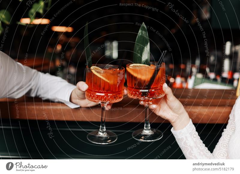 hands of woman and man are clinking, cheers with glasses of Spritz cocktail. Couple celebrating wedding, anniversary with Aperol spritz cocktails, with orange and greens Refreshing alcoholic drink