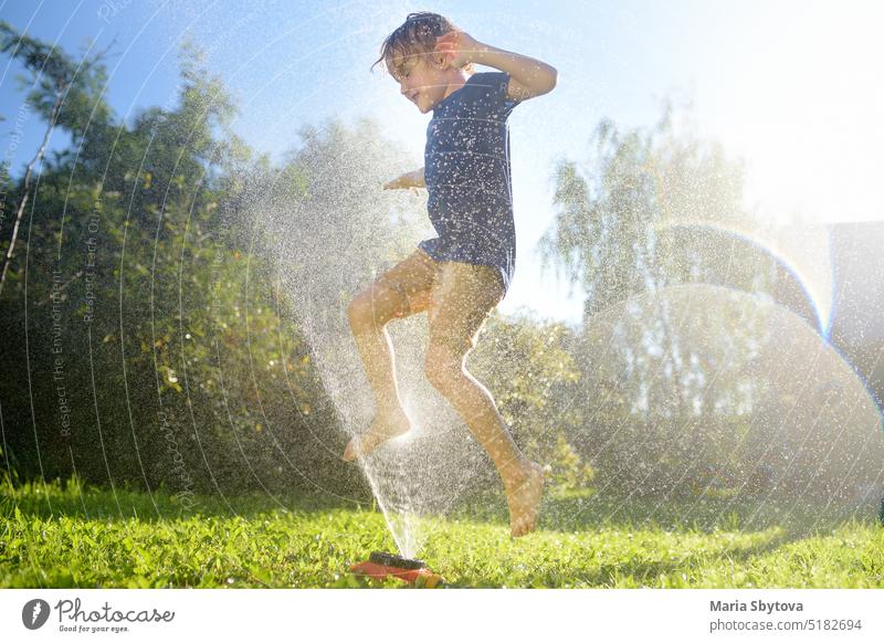 Funny little boy playing with garden sprinkler in sunny backyard. Preschooler child laughing, jumping and having fun with spray of water. Summer holidays for kids in the village