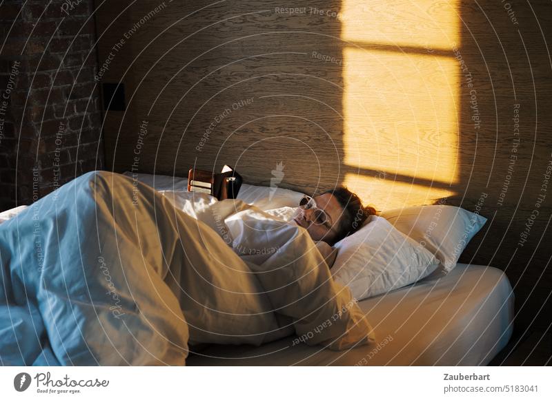 Woman lies in comfortable white feather bed and reads in cell phone, light shines from window into room Reading Bed Cellphone Cozy Quilt White Hotel Window Sun