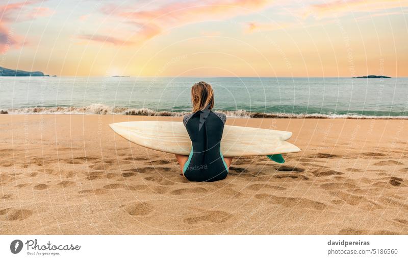 Surfer woman with wetsuit and surfboard sitting on the sand looking at the sea unrecognizable young surfer back beach people 20s outdoors twenties anonymous