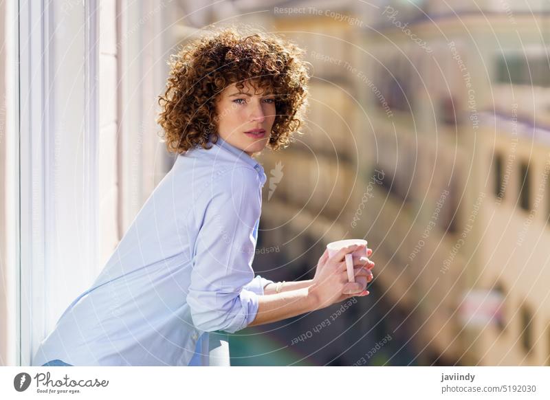 Curly haired woman drinking coffee on balcony morning breakfast teatime coffee break lean on railing female adult brown hair free time apartment cup leisure
