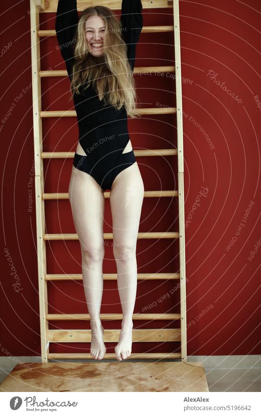 a young blonde slim woman in black bodysuit hangs on the wall bars over a gym mat in front of a red wall Woman youthful Slim Athletic pretty Smiling fit