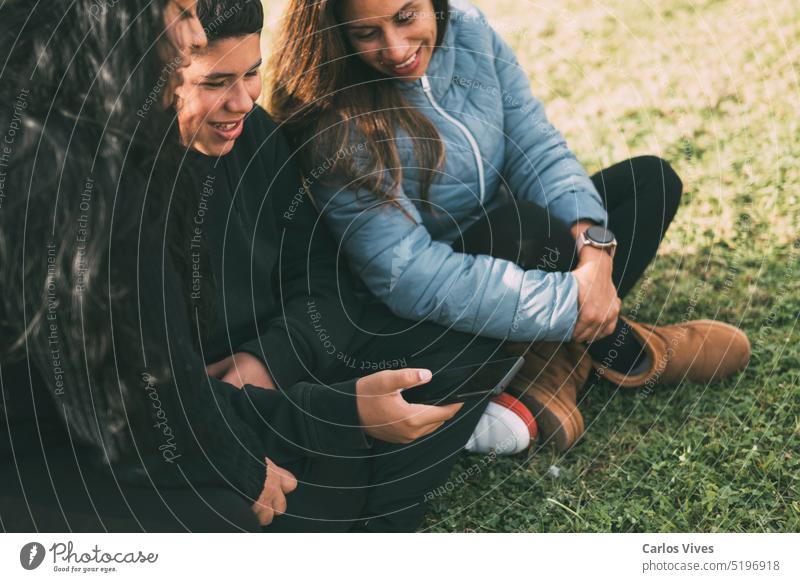 Hispanic male teenager holding smartphone looking away while sitting on grass with Hispanic mother and sister in park on sunny day Hispanic culture