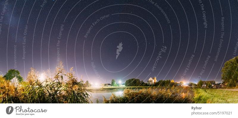 Panorama Of Evaporation Over River Lake Near Houses In Village. Night Starry Sky Above Lake River With Bright Stars And Meteoric Track Trail. Glowing Stars Above Summer Nature