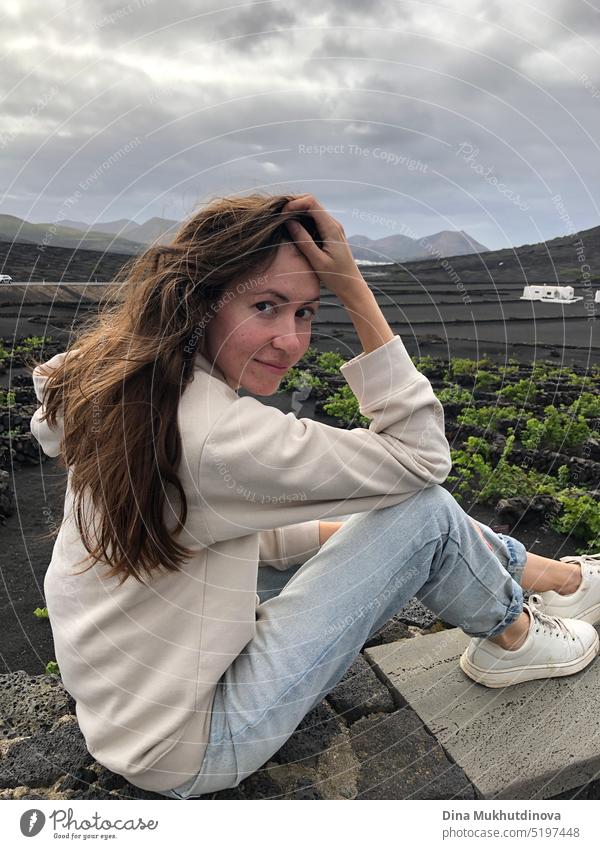 Female tourist in Lanzarote, Canary Islands. Young caucasian woman in jeans and hoodie sitting on the bench posing for a photo at the winery. Wine tourism.
