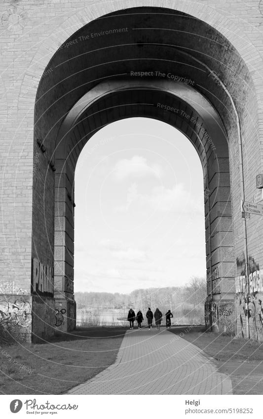 under the arch of a viaduct are 5 unrecognizable, seemingly tiny people, 4 on foot, one by bicycle Manmade structures Architecture Silhouette Pedestrian