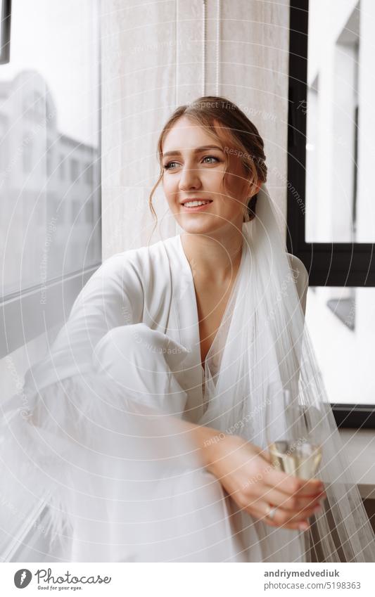 Bride s morning. Bride drinking champagne in the peignoir. young woman is sitting on a large window in a hotel room in bathroom. Beautiful girl in white wedding robe. wedding day