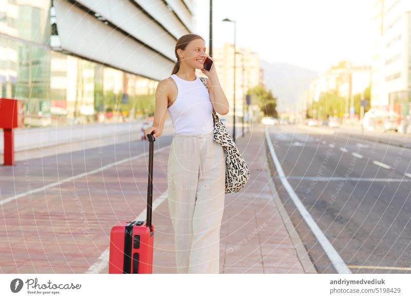 young beautiful woman with red suitcase calling smartphone while waiting for a taxi cab in the city. stylish smiling girl tourist on summer holiday vacation. new trip. copy space