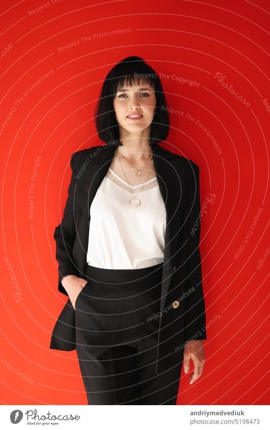 Beautiful female brunette business woman CEO in a black suit, isolated on red background, standing confidently with arms in pockets. Females in Business.