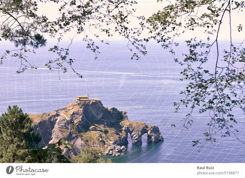 Hermitage of San Juan de Gaztelugatxe on a sunny day. calm sea, rocky formation, holes, frame of branches and trees sunlight island ocean vacations horizontal