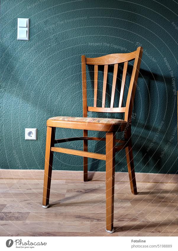 The empty chair in front of a petrol-colored wall on laminate floor casts its shadow on the wall. The socket and light switch glow white. Wall (building)