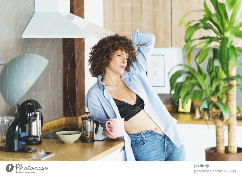 Hot woman in black lingerie shirt and jeans with cup of tea touch hair touch head teatime hot drink unbutton kitchen female adult curly hair free time lean on