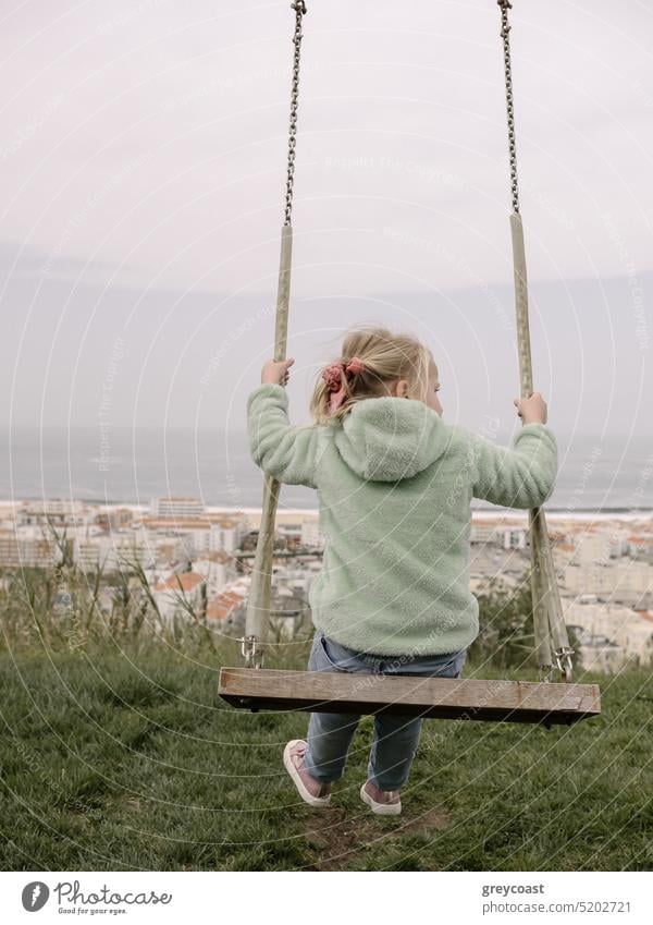 Swinging on Wooden Swing with Ocean and City View girl swinging vertical scandic scandinavian back from back blonde wooden hill back view ocean child outdoors