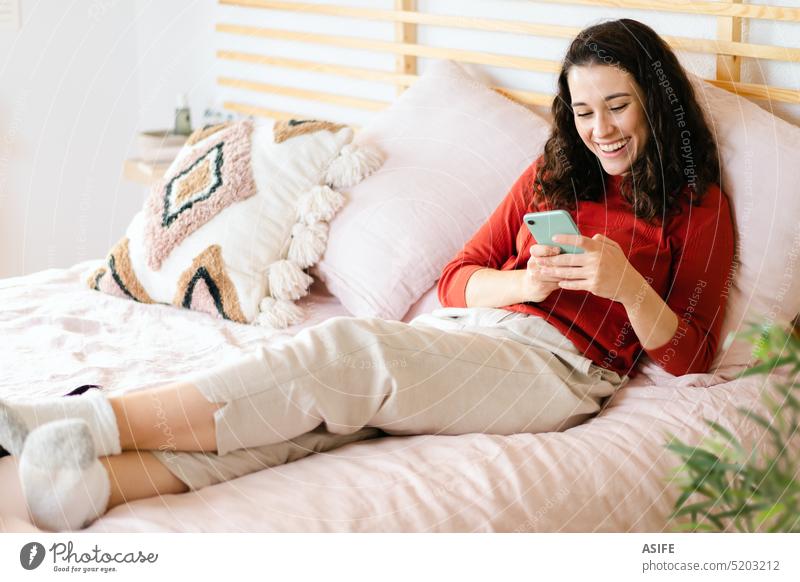 Woman relaxing and having fun with a smartphone on the bed young woman mobile phone technology laughing happy smiling relaxed comfortable lying leisure cozy