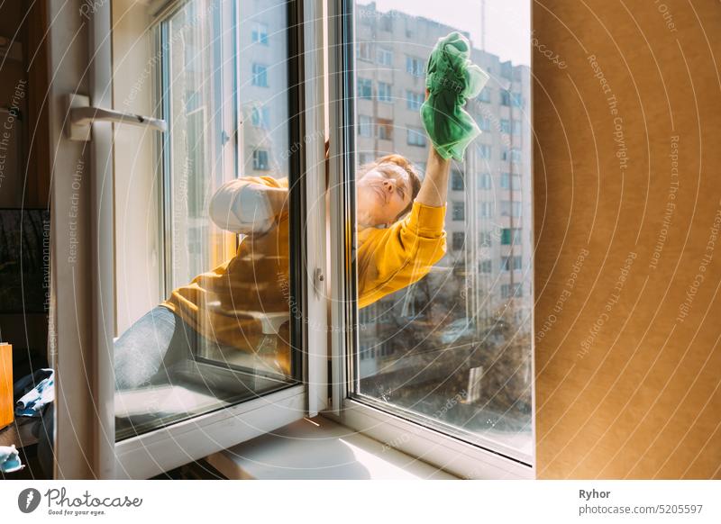 Caucasian Woman Of Fifty In A Yellow Sweater And Jeans Washes A Dusty Window In Apartment. A 50 Year Old Woman Cleans Windows From Stains Using Rag. Elderly Woman Is Cleaning House, Doing Household Chores
