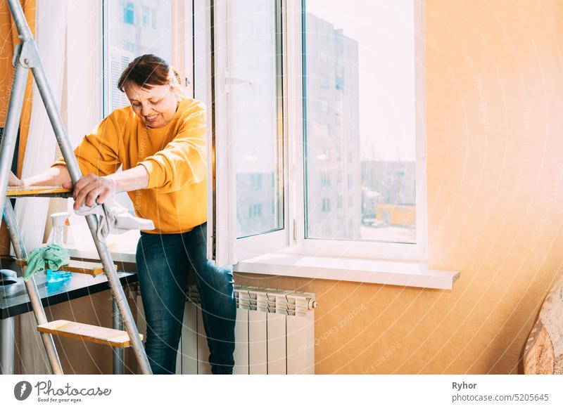 Smiling Caucasian Woman Of Fifty In Yellow Sweater And Jeans Washes Dusty Window In Apartment. 50 Year Old Woman Cleans Windows From Stains Using Rag And Spray Cleaner. Elderly Woman Is Cleaning House, Doing Household Chores. Happy Woman, Satisfied With...