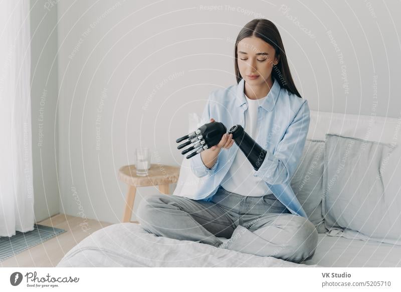 Disabled young girl disassembles bionic prosthetic arm, sitting on bed at home. Medical technologies prosthesis disabled artificial limb grasp grip hand touch