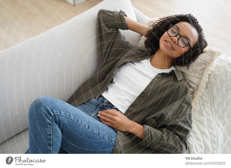 Relaxed serene young mixed race girl resting lying on comfortable sofa, enjoy afternoon nap at home couch living room sleep relax glasses wellness doze calm