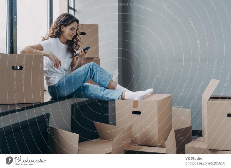 Girl holding phone sitting on windowsill with boxes selects moving service, planning home renovation female relocation smartphone app cardboard box removal