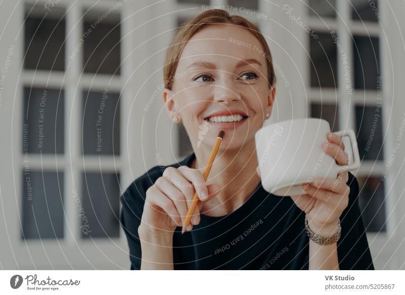 Successful mid adult businesswoman is drinking tea, smiling and dreaming at workplace . coffee cup pencil hand portrait career success businesswoman dreaming