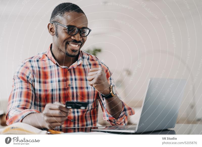 African american man holding credit card uses online banking service at laptop, makes yes gesture african american success money cashback e commerce purchase
