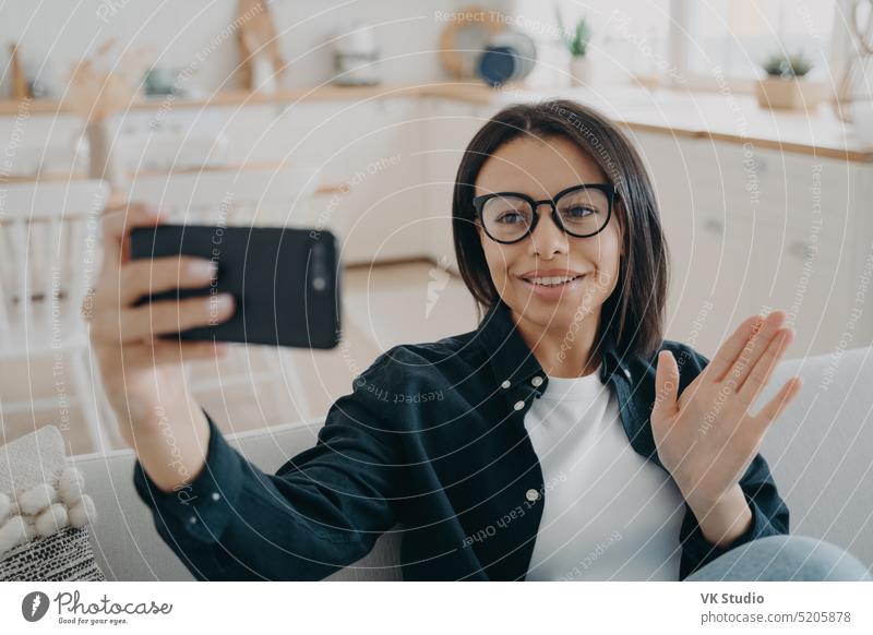 Female in glasses answers video call holding smartphone, greeting waving hand hello, sitting at home friendly lifestyle application woman living room day