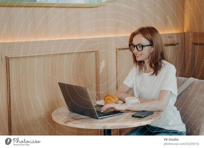 Happy smiling woman in earphones using laptop while sitting at cafe freelancer work distant education computer internet young businesswoman breakfast croissant