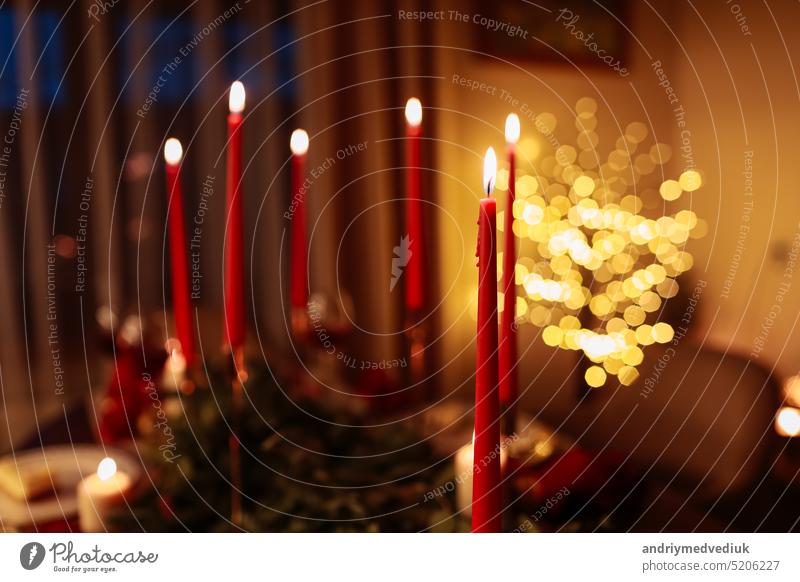 Burning long red candles on festive table for celebration Christmas and New Year holidays in living room on xmas tree with garlands background. Dining table decorated fir branches and xmas baubles