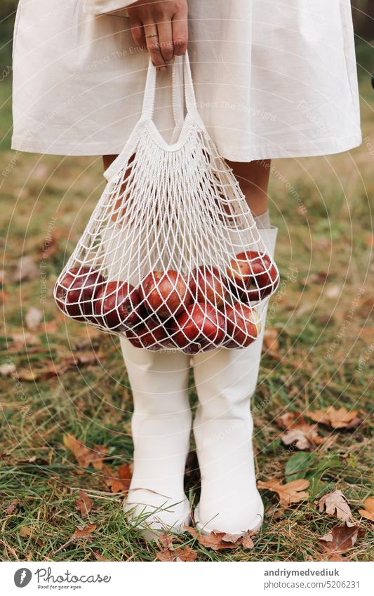 String bag with red apples. View of female legs in white boots walking through the autumn garden with eco bag in hands. Zero waste concept. No plastic life. Healthy fitness lifestyle. Recycling waste