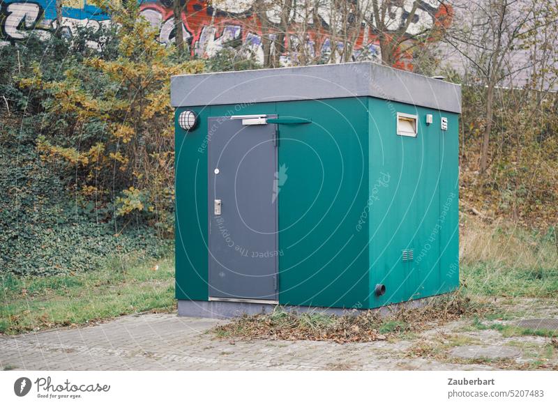 Cubic cottage in turquoise green stands in front of bushes and concrete wall Small square Sharp-edged Turquoise Green Toilet Miniature door toilet house