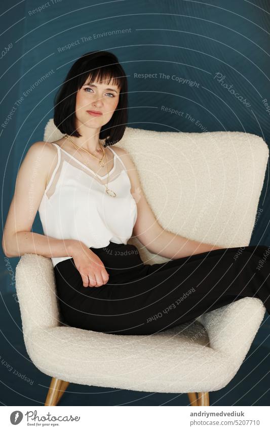 attractive business woman sits in comfy white chair. Smiling brunette lady with short hair wears black office trousers and white blouse, high-heels isolated on blue green background. copy space.