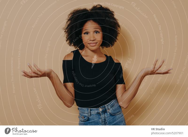 Confused unaware Afro woman with wavy curly hair, raises hands in bewilderment beauty not sure beige decision make cannot haircut individual indignant