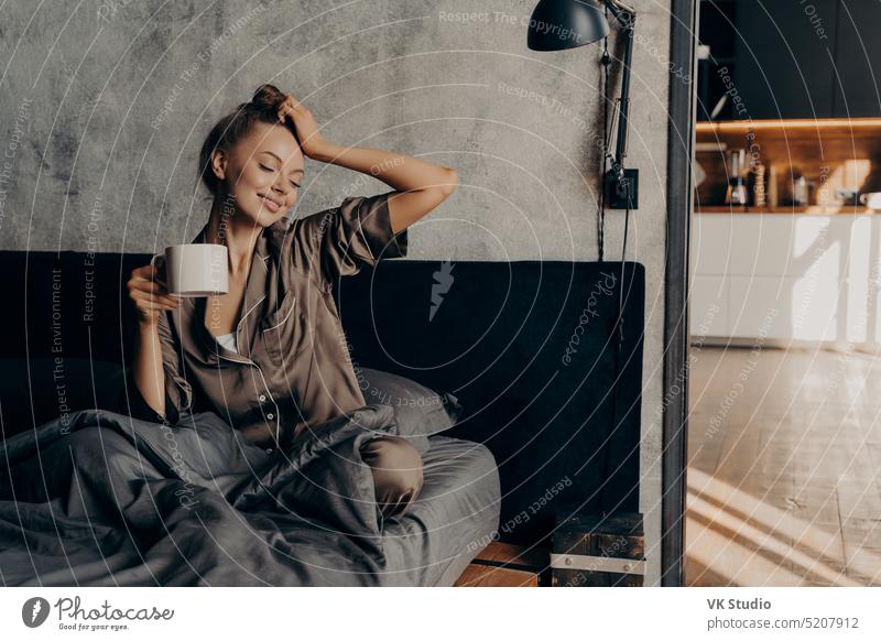 Portrait of young relaxed woman in stylish home suit waking up with cup of coffee in bed portrait cozy bedroom stretching from night sleep enjoying morning