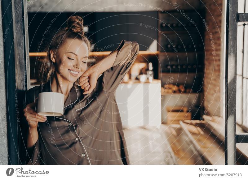Happy beautiful brunette female stratching with cup of coffee in her hand while standing in kitchen young pretty woman morning mug stretching bedroom smiling