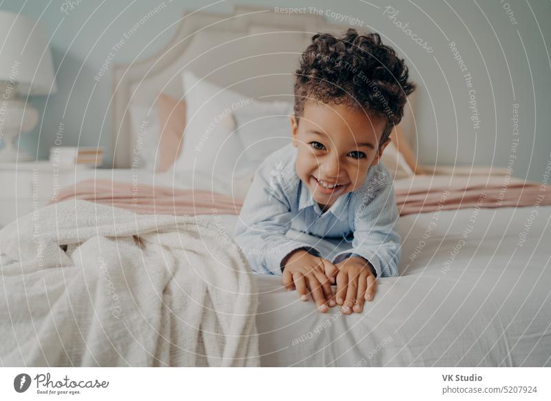 Happy curly afro american boy laying on bed and smiling at camera cute little child mixed race african portrait happy mulatto cozy relaxing fun playing home