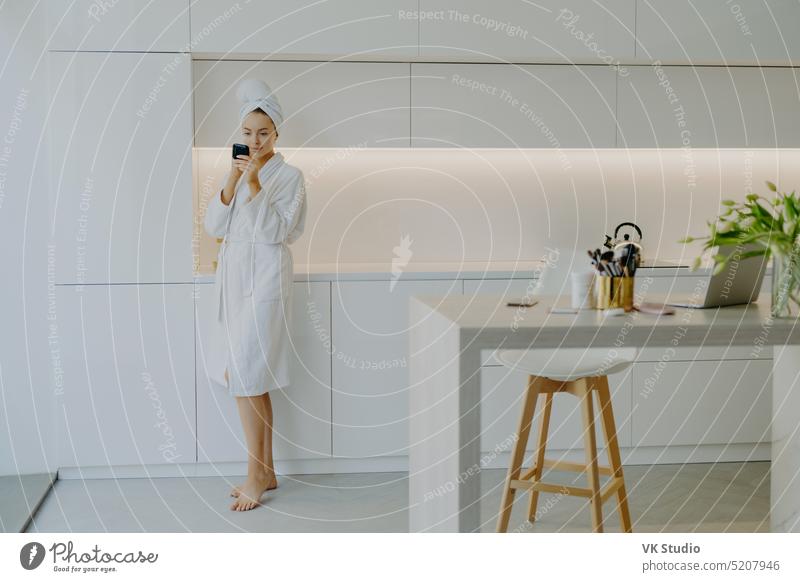 Full length shot of young beautiful woman dressed in white bathrobe applies face cream looks in mirror takes care of her skin poses near kitchen furniture stands bare feet. People beauty facial care
